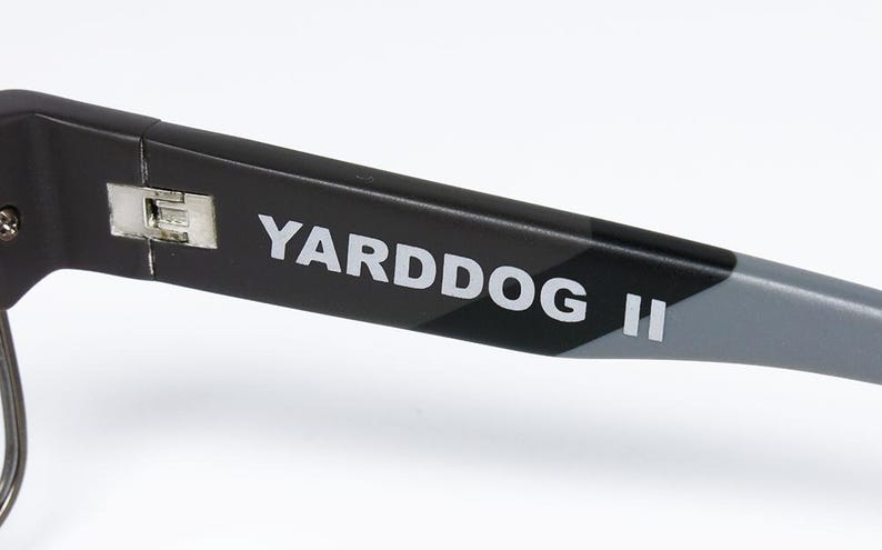 May include: Close-up of the arm of a pair of black sunglasses with the text "YARDDOG II" in white. The arm has a camouflage pattern in black and gray.