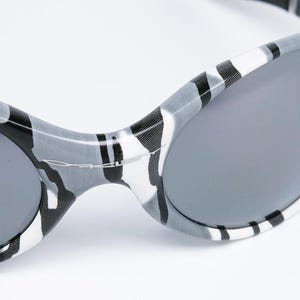 May include: Close-up of sunglasses with a gray and white camouflage pattern on the frames. The lenses are a solid gray color. The sunglasses have a sporty, curved design.