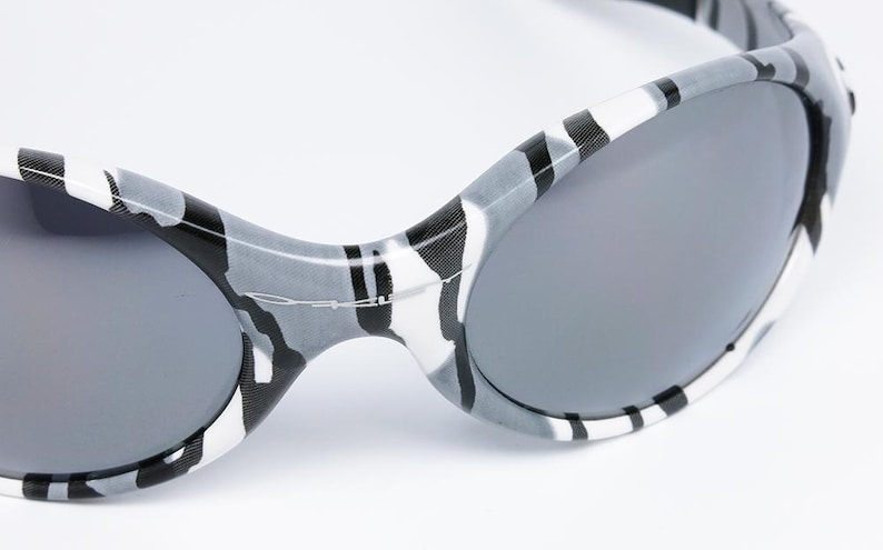 May include: Close-up of sunglasses with a gray and white camouflage pattern on the frames. The lenses are a solid gray color. The sunglasses have a sporty, curved design.