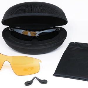 May include: Black sunglasses with a black case, extra orange lenses, a black pouch, and a nose piece. The sunglasses are in an open, black, oval-shaped case. The extra lenses are orange.