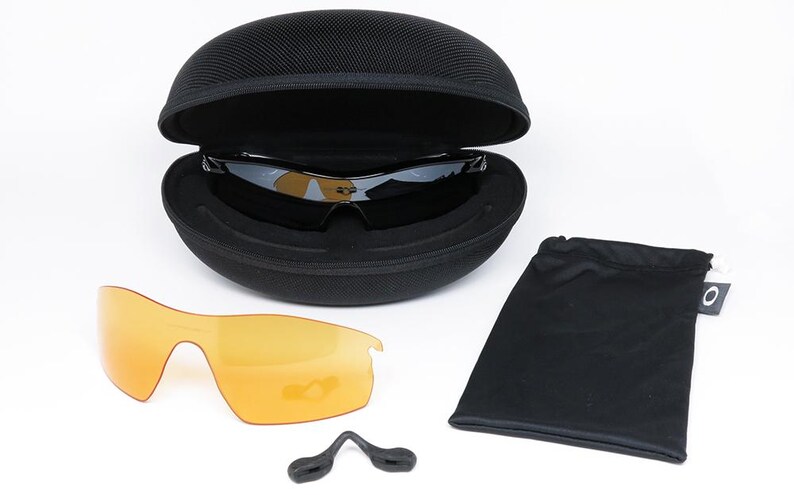 May include: Black sunglasses with a black case, extra orange lenses, a black pouch, and a nose piece. The sunglasses are in an open, black, oval-shaped case. The extra lenses are orange.