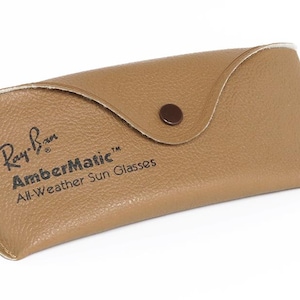 Puede incluir: Un estuche de gafas de sol de cuero marr&oacute;n claro con cierre de bot&oacute;n a presi&oacute;n. El estuche tiene el logotipo de Ray-Ban y las palabras "AmberMatic All-Weather Sun Glasses" impresas. El estuche est&aacute; dise&ntilde;ado para proteger las gafas de sol.