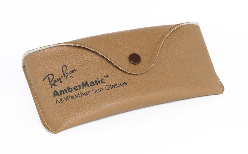 Puede incluir: Un estuche de gafas de sol de cuero marr&oacute;n claro con cierre de bot&oacute;n a presi&oacute;n. El estuche tiene el logotipo de Ray-Ban y las palabras "AmberMatic All-Weather Sun Glasses" impresas. El estuche est&aacute; dise&ntilde;ado para proteger las gafas de sol.