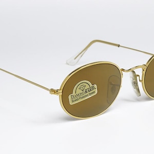 Pu&ograve; includere: Occhiali da sole ovali con montatura dorata e lenti marroni. Gli occhiali hanno una montatura dorata sottile e un logo Ray-Ban sulla lente. Le lenti hanno un adesivo con la scritta "Diamond Hard Scratch Protection System".