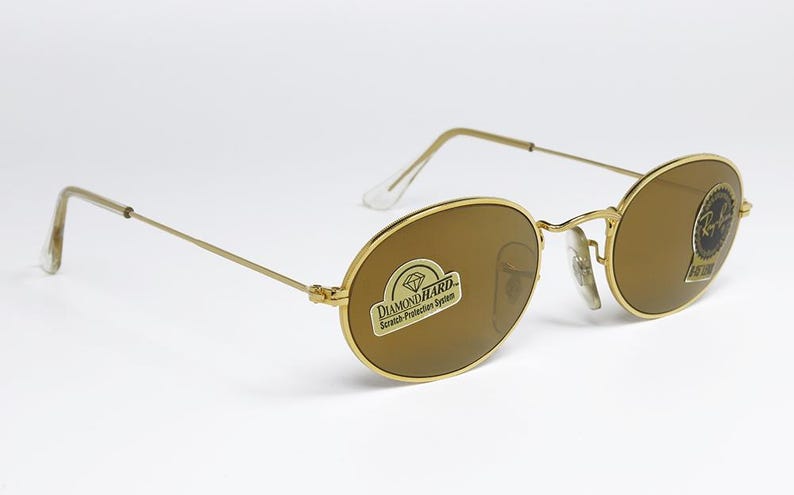 Pu&ograve; includere: Occhiali da sole ovali con montatura dorata e lenti marroni. Gli occhiali hanno una montatura dorata sottile e un logo Ray-Ban sulla lente. Le lenti hanno un adesivo con la scritta "Diamond Hard Scratch Protection System".