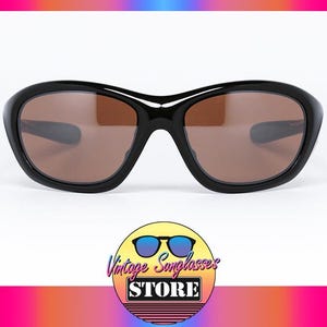 K&ouml;nnte beinhalten: Schwarze Sonnenbrille mit braunen Gl&auml;sern. Die Sonnenbrille hat einen abgerundeten, dicken Rahmen. Das Bild enth&auml;lt ein Logo f&uuml;r einen Vintage-Sonnenbrillenladen.