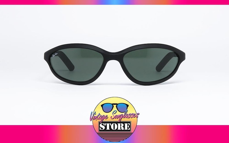 Pode incluir: &Oacute;culos de sol Ray-Ban pretos com arma&ccedil;&atilde;o oval e lentes verde escuro. Os &oacute;culos de sol t&ecirc;m um acabamento fosco e um logotipo Ray-Ban sutil na haste. A imagem est&aacute; em um fundo branco.