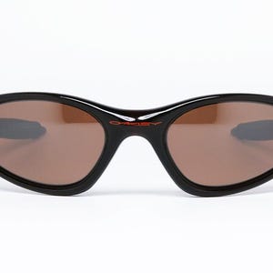 Pode incluir: &Oacute;culos de sol pretos com lentes castanhas. Os &oacute;culos t&ecirc;m uma forma oval curvada com detalhes vermelhos nas hastes. O nome da marca "Oakley" est&aacute; impresso em vermelho na arma&ccedil;&atilde;o.
