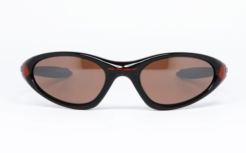 Pode incluir: &Oacute;culos de sol pretos com lentes castanhas. Os &oacute;culos t&ecirc;m uma forma oval curvada com detalhes vermelhos nas hastes. O nome da marca "Oakley" est&aacute; impresso em vermelho na arma&ccedil;&atilde;o.