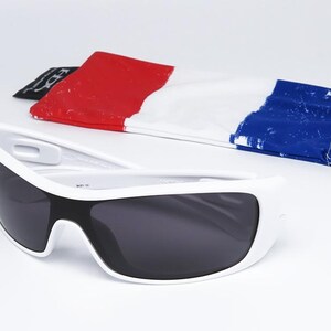 Op de afbeelding: Witte zonnebril met donkere glazen en een etui in de kleuren van de Franse vlag: rood, wit en blauw. De zonnebril heeft een sportief, modern design.