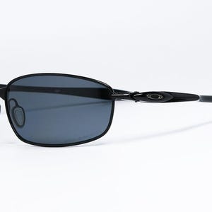 Puede incluir: Gafas de sol negras con montura rectangular y lentes oscuras. Las gafas tienen un dise&ntilde;o elegante con una montura fina y patillas curvas. El logotipo de Oakley es visible en la patilla.