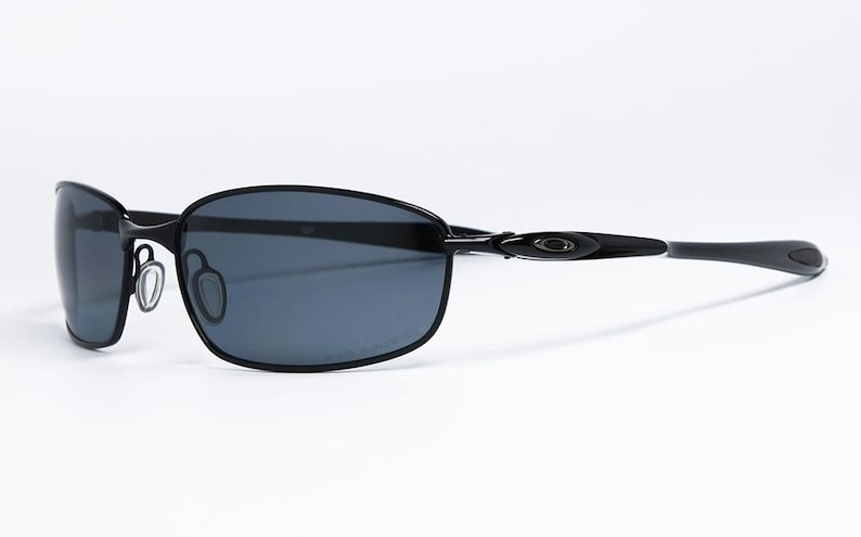 Puede incluir: Gafas de sol negras con montura rectangular y lentes oscuras. Las gafas tienen un dise&ntilde;o elegante con una montura fina y patillas curvas. El logotipo de Oakley es visible en la patilla.