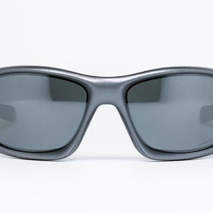 May include: Gray sunglasses with dark lenses. The sunglasses have a curved frame and a small red accent on each side. The lenses are a dark gray color, and the frame is a matte gray finish. The sunglasses are designed for sports and outdoor activities.