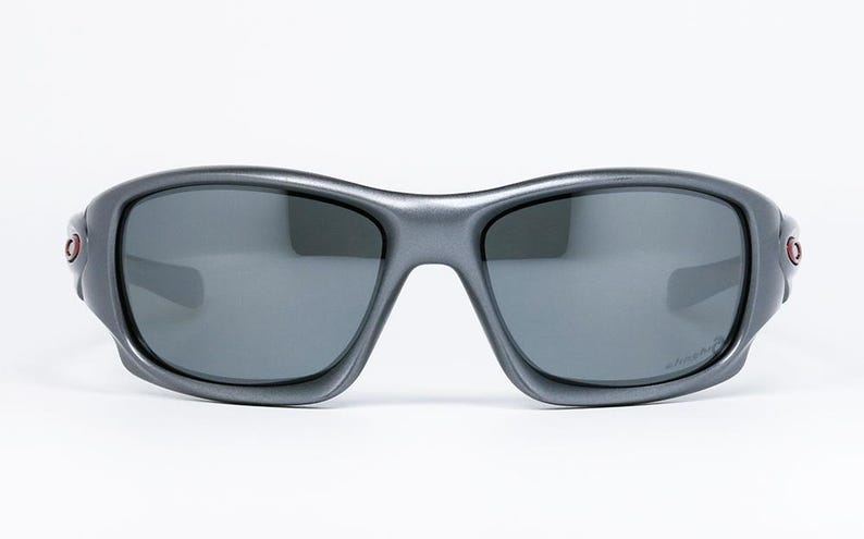 May include: Gray sunglasses with dark lenses. The sunglasses have a curved frame and a small red accent on each side. The lenses are a dark gray color, and the frame is a matte gray finish. The sunglasses are designed for sports and outdoor activities.