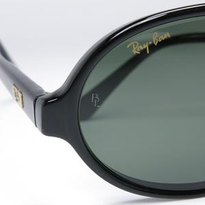 May include: Close-up of black Ray-Ban sunglasses. The sunglasses have a glossy black frame and dark green lenses. The Ray-Ban logo is in gold lettering on the upper left lens. The sunglasses are a classic style.