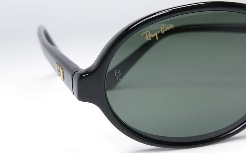 May include: Close-up of black Ray-Ban sunglasses. The sunglasses have a glossy black frame and dark green lenses. The Ray-Ban logo is in gold lettering on the upper left lens. The sunglasses are a classic style.