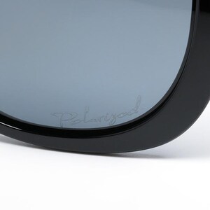 Puede incluir: Primer plano de gafas de sol negras con lentes polarizadas grises. La palabra "Polarized" est&aacute; escrita en una fuente de guion en la lente. Las gafas de sol tienen una montura gruesa y redondeada.