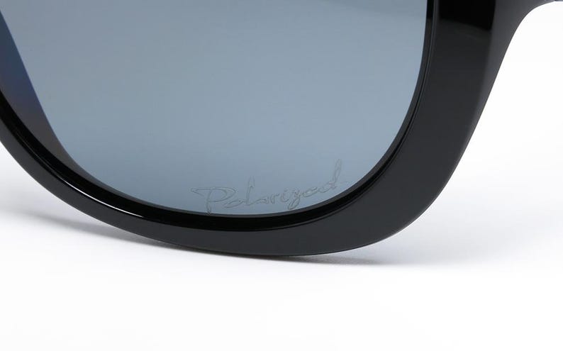 Puede incluir: Primer plano de gafas de sol negras con lentes polarizadas grises. La palabra "Polarized" est&aacute; escrita en una fuente de guion en la lente. Las gafas de sol tienen una montura gruesa y redondeada.