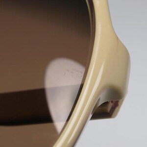 May include: Close-up of sunglasses with a light tan frame and brown lenses. The sunglasses have a sleek, modern design. The lenses reflect light, creating a bright, contrasting effect. The frame is smooth and glossy.