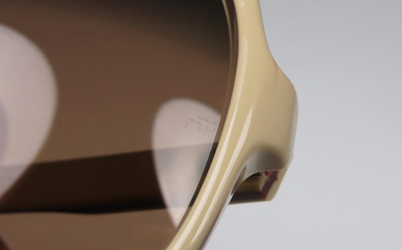 May include: Close-up of sunglasses with a light tan frame and brown lenses. The sunglasses have a sleek, modern design. The lenses reflect light, creating a bright, contrasting effect. The frame is smooth and glossy.