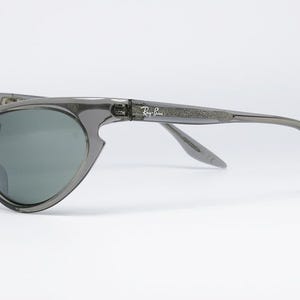 May include: Gray sunglasses with a wraparound design. The lenses are a dark gray color, and the frames are a translucent gray. The Ray-Ban logo is visible on the temple. These sunglasses are a stylish accessory.