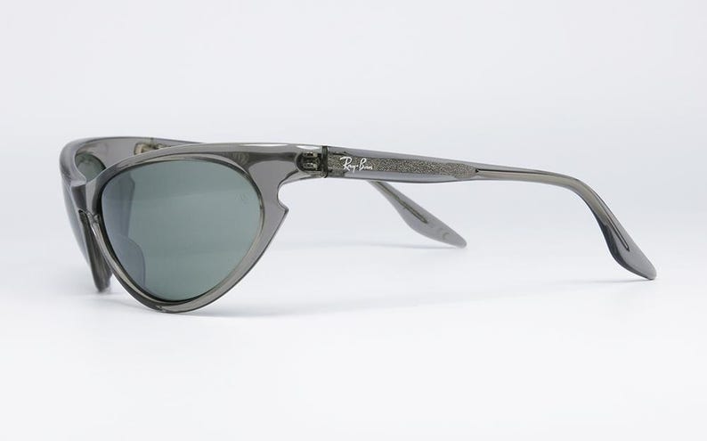 May include: Gray sunglasses with a wraparound design. The lenses are a dark gray color, and the frames are a translucent gray. The Ray-Ban logo is visible on the temple. These sunglasses are a stylish accessory.
