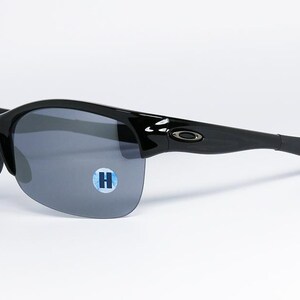 May include: Black sunglasses with gray lenses. The sunglasses have a sleek, curved design with a glossy finish. The lenses are a dark gray color, and the frame is black. A small blue and white sticker with the letter "H" is on the lens.