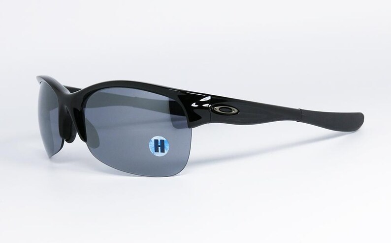 May include: Black sunglasses with gray lenses. The sunglasses have a sleek, curved design with a glossy finish. The lenses are a dark gray color, and the frame is black. A small blue and white sticker with the letter "H" is on the lens.