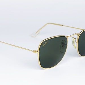 May include: Square sunglasses with a gold frame and green lenses. The sunglasses have a Ray-Ban logo on the right lens.