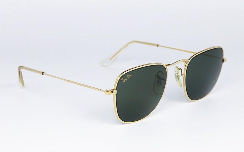 May include: Square sunglasses with a gold frame and green lenses. The sunglasses have a Ray-Ban logo on the right lens.