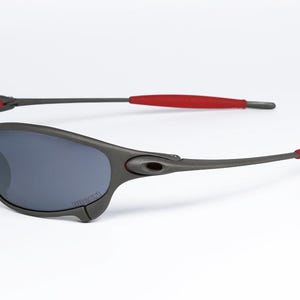 May include: Gray sunglasses with dark lenses and red accents on the temples. The sporty eyewear features a wraparound design and a sleek, modern aesthetic. The sunglasses are set against a white background.