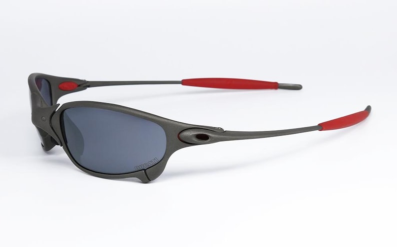 May include: Gray sunglasses with dark lenses and red accents on the temples. The sporty eyewear features a wraparound design and a sleek, modern aesthetic. The sunglasses are set against a white background.