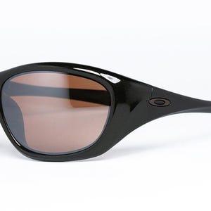 K&ouml;nnte beinhalten: Schwarze Sonnenbrille mit braun get&ouml;nten Gl&auml;sern. Die Sonnenbrille hat ein geschwungenes, umlaufendes Design mit gl&auml;nzendem Finish. Das Oakley-Logo ist auf dem B&uuml;gel sichtbar. Diese Sonnenbrille ist f&uuml;r Outdoor-Aktivit&auml;ten konzipiert.