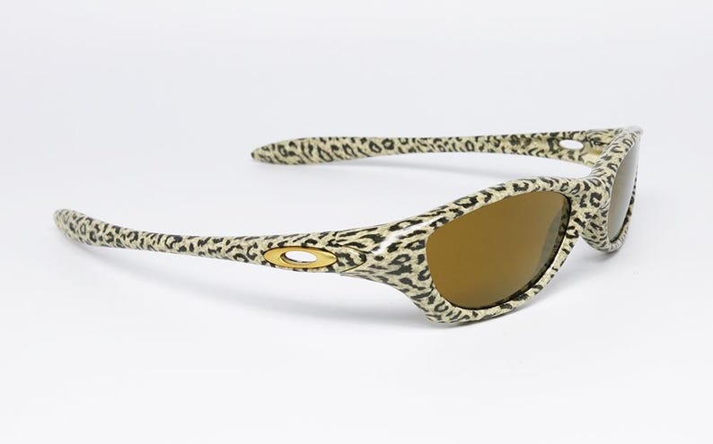 May include: Leopard print sunglasses with brown lenses. The frames feature a tan and black spotted pattern. The Oakley logo is visible on the temples. These sunglasses are a stylish accessory for eye protection.