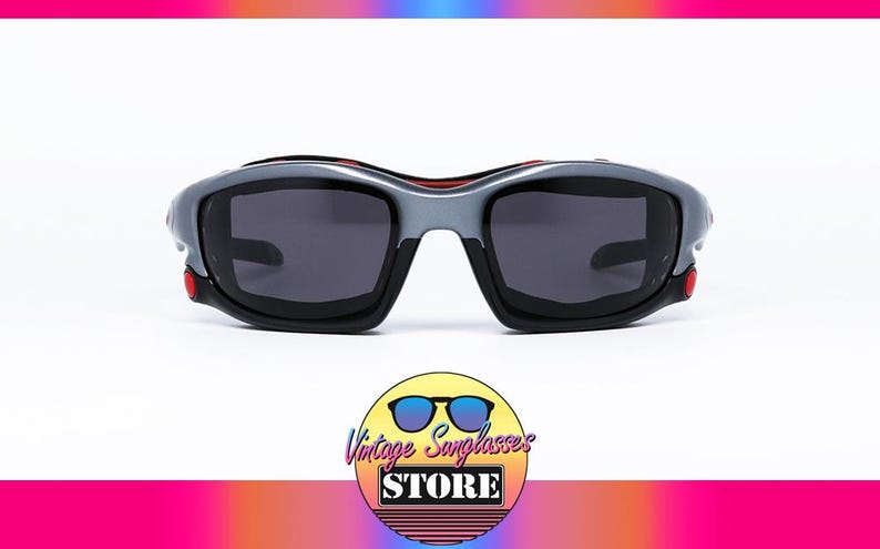 Puede incluir: Gafas de sol envolventes grises y negras con detalles rojos. Las lentes son de color gris oscuro.
