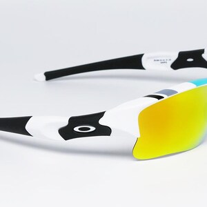 May include: White and black sunglasses with yellow mirrored lenses. The sunglasses have a sporty design with a white frame, black arms, and a black logo on the side. The lenses are a bright yellow color.