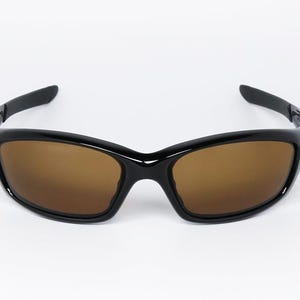 May include: Black sunglasses with brown lenses. The sunglasses have a glossy black frame and a curved design. The lenses are a warm brown colour, providing a contrast to the black frame. The arms of the sunglasses are also black.
