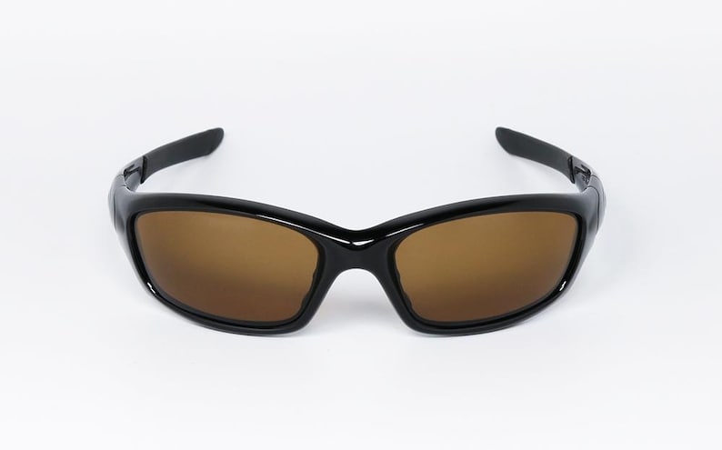 May include: Black sunglasses with brown lenses. The sunglasses have a glossy black frame and a curved design. The lenses are a warm brown colour, providing a contrast to the black frame. The arms of the sunglasses are also black.