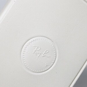 Puede incluir: Primer plano de una superficie blanca texturizada con un borde redondeado. Se ve un logotipo circular en relieve con el texto "Ray-Ban". El dise&ntilde;o sugiere un producto asociado con la marca Ray-Ban.