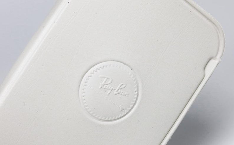 Puede incluir: Primer plano de una superficie blanca texturizada con un borde redondeado. Se ve un logotipo circular en relieve con el texto "Ray-Ban". El dise&ntilde;o sugiere un producto asociado con la marca Ray-Ban.
