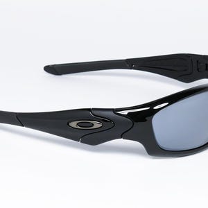 May include: Black sunglasses with gray lenses. The sunglasses have a sleek, modern design with curved frames and arms. The Oakley logo is visible on the temples. The lenses are a reflective gray color.