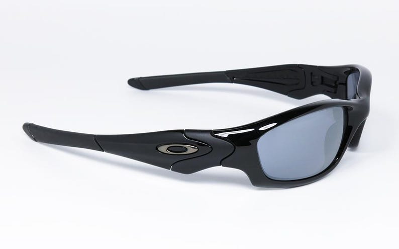 May include: Black sunglasses with gray lenses. The sunglasses have a sleek, modern design with curved frames and arms. The Oakley logo is visible on the temples. The lenses are a reflective gray color.