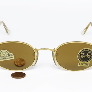 Pu&ograve; includere: Occhiali da sole ovali con montatura dorata e lenti marroni. Gli occhiali hanno un ponte e aste dorate e le lenti sono leggermente colorate. Le lenti hanno adesivi con la scritta "Diamond Hard" e "Ray-Ban B-15 Lens". Una moneta &egrave; posta sulla lente.