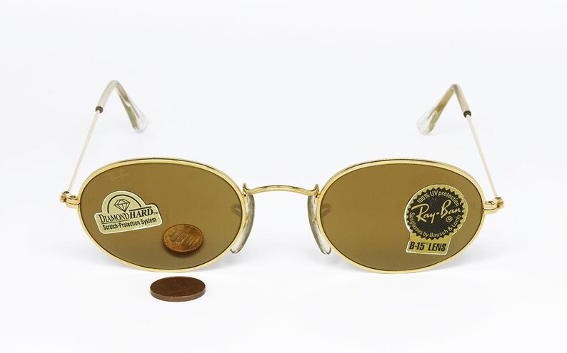 Pu&ograve; includere: Occhiali da sole ovali con montatura dorata e lenti marroni. Gli occhiali hanno un ponte e aste dorate e le lenti sono leggermente colorate. Le lenti hanno adesivi con la scritta "Diamond Hard" e "Ray-Ban B-15 Lens". Una moneta &egrave; posta sulla lente.