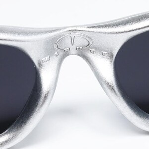 May include: Close-up of silver sunglasses with dark lenses. The sunglasses have a distinctive design with a bridge and a logo detail. The lenses are a dark color, and the frame has a metallic finish. The sunglasses are a fashion accessory.