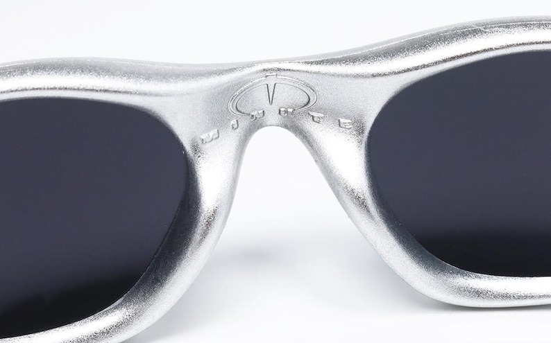 May include: Close-up of silver sunglasses with dark lenses. The sunglasses have a distinctive design with a bridge and a logo detail. The lenses are a dark color, and the frame has a metallic finish. The sunglasses are a fashion accessory.