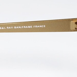 Puede incluir: Primer plano de una patilla de gafas de sol doradas. El texto "B&L RAY-BAN/FRAME FRANCE" est&aacute; impreso en blanco. La bisagra y el borde de la lente verde oscuro son visibles sobre un fondo blanco.