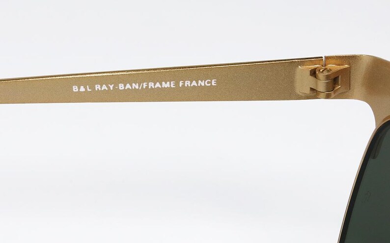 Puede incluir: Primer plano de una patilla de gafas de sol doradas. El texto "B&L RAY-BAN/FRAME FRANCE" est&aacute; impreso en blanco. La bisagra y el borde de la lente verde oscuro son visibles sobre un fondo blanco.