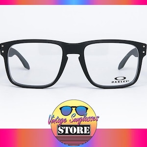 Puede incluir: Gafas Oakley negras con montura cuadrada y lentes transparentes. El logotipo de Oakley es visible en la lente derecha. Las gafas están sobre un fondo blanco con un degradado de color en la parte superior e inferior.