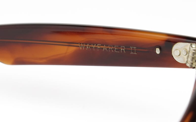 Peut inclure: Gros plan sur la monture en &eacute;caille brune d'une paire de lunettes de soleil Wayfarer II. La monture est dot&eacute;e d'une charni&egrave;re argent&eacute;e et le texte "WAYFARER II" est grav&eacute; sur le c&ocirc;t&eacute;.